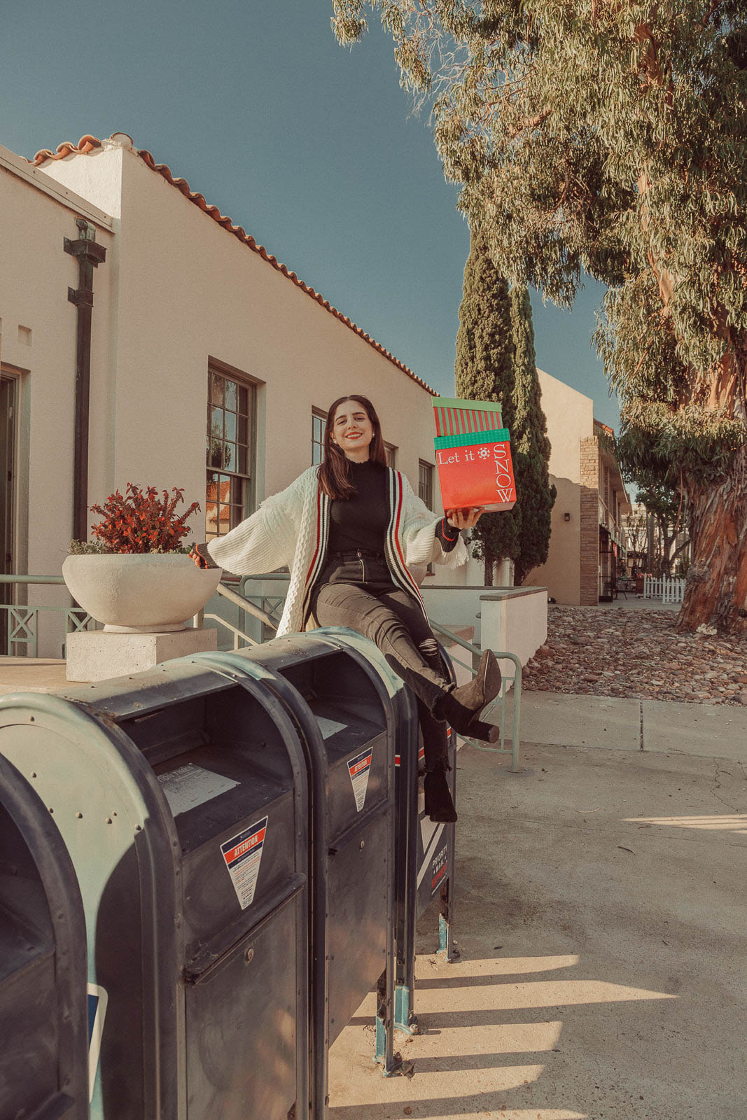 Woman wearing white sweater holding Christmas gifts sitting on mailbox - 5 Creative Christmas Photoshoot Ideas to Try This Season