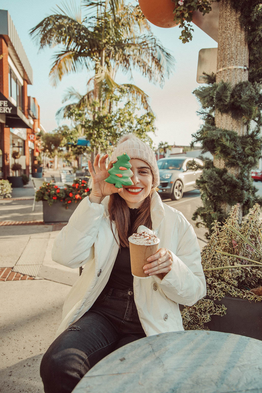 Woman wearing white knit beanie and  puffer jacket holding Christmas tree shaped sugar cookie to her eye and holding a hot chocolate cup while sitting- 5 Creative Christmas Photoshoot Ideas to Try This Season