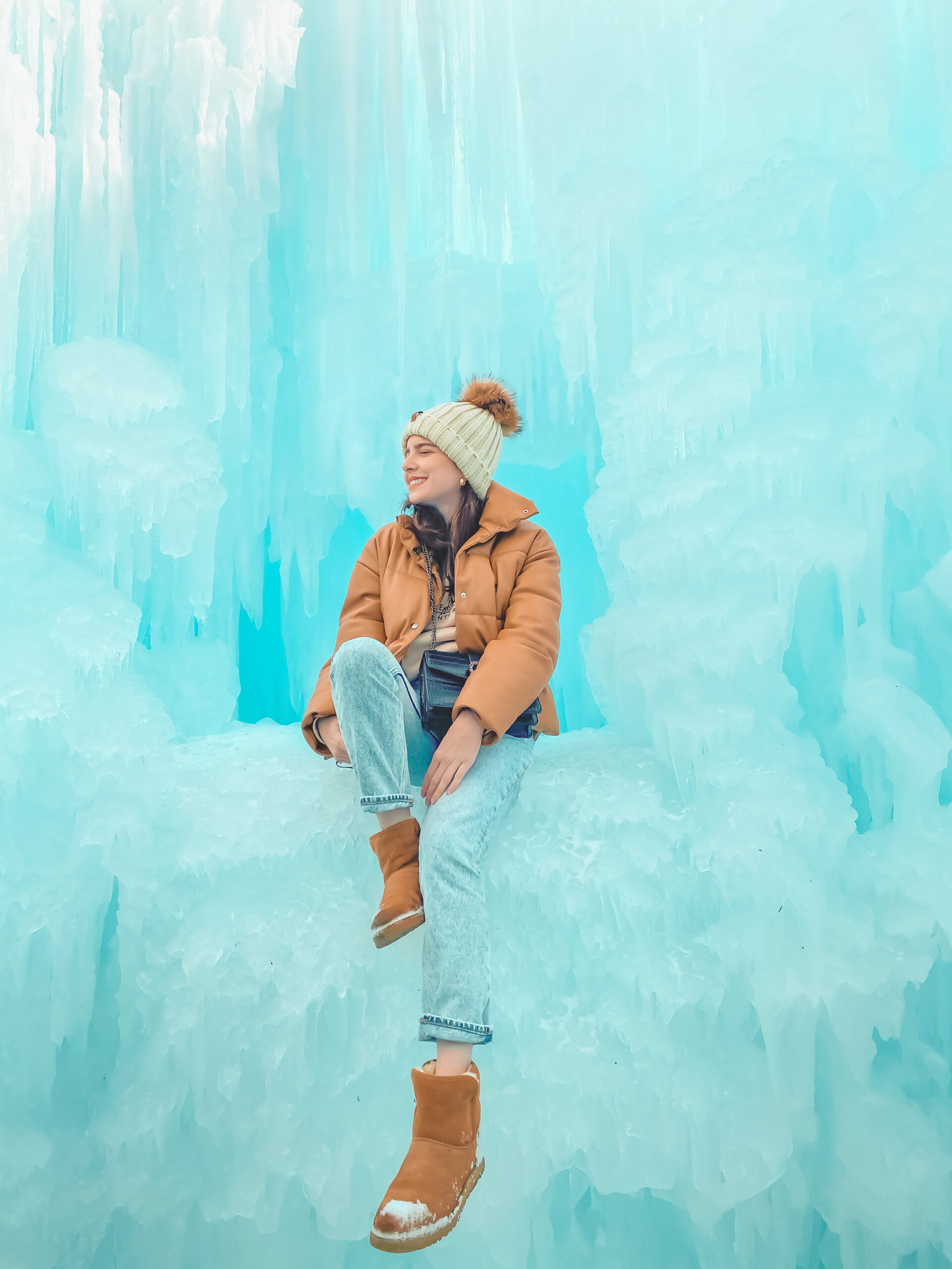 Tips for Visiting the Ice Castles in Midway, UT, woman wearing pompom beanie, tan puffer coat, and Uggs boots sitting on ice - Palm Trees and Pellegrino Travel Tips