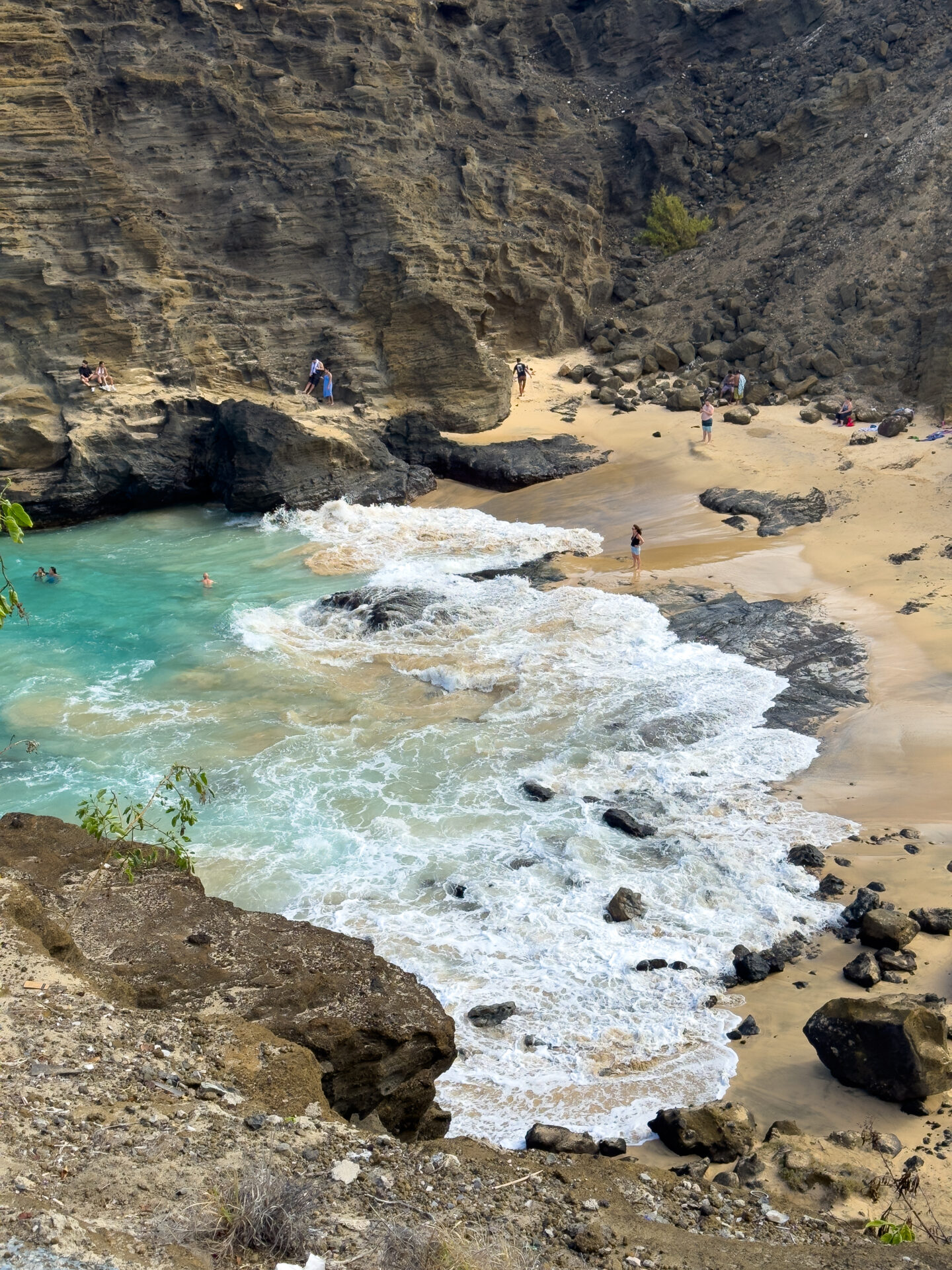 The shore of Halona Beach Cove with people swimming, standing on the sand, and sitting on the rocks.